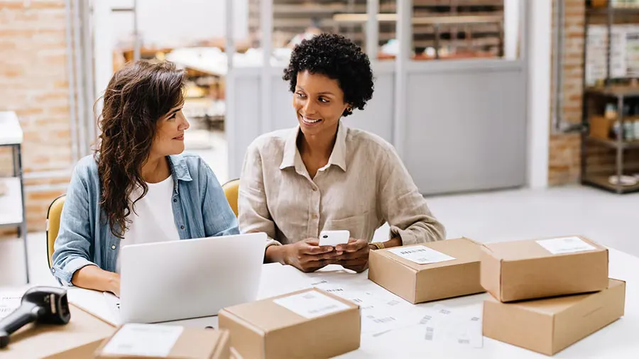 Two business owners managing ecommerce orders with laptops, phones, and shipping boxes.